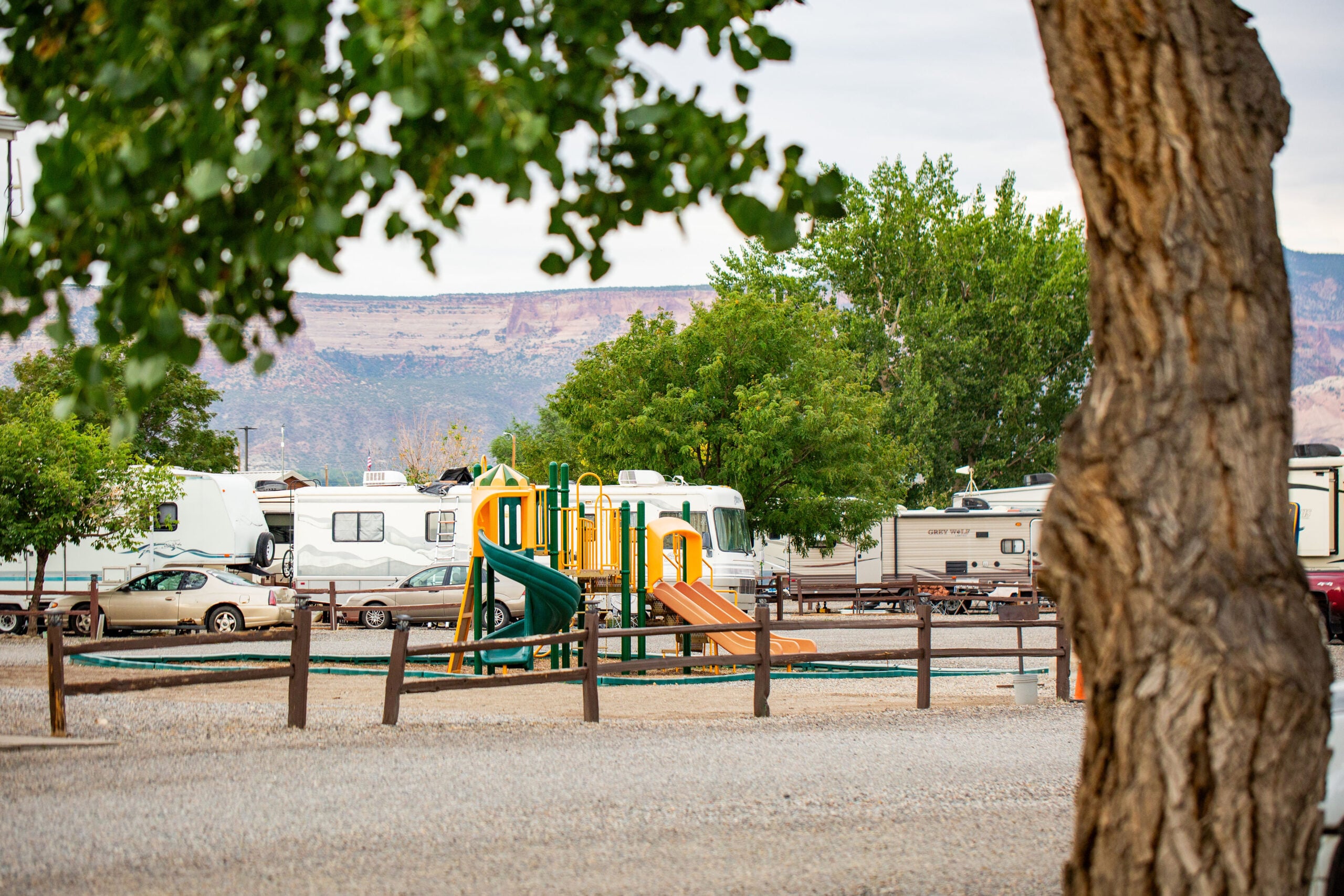 A playground with slides and climbing structures is set among RVs in a gravel lot, with trees and a mountain in the background.