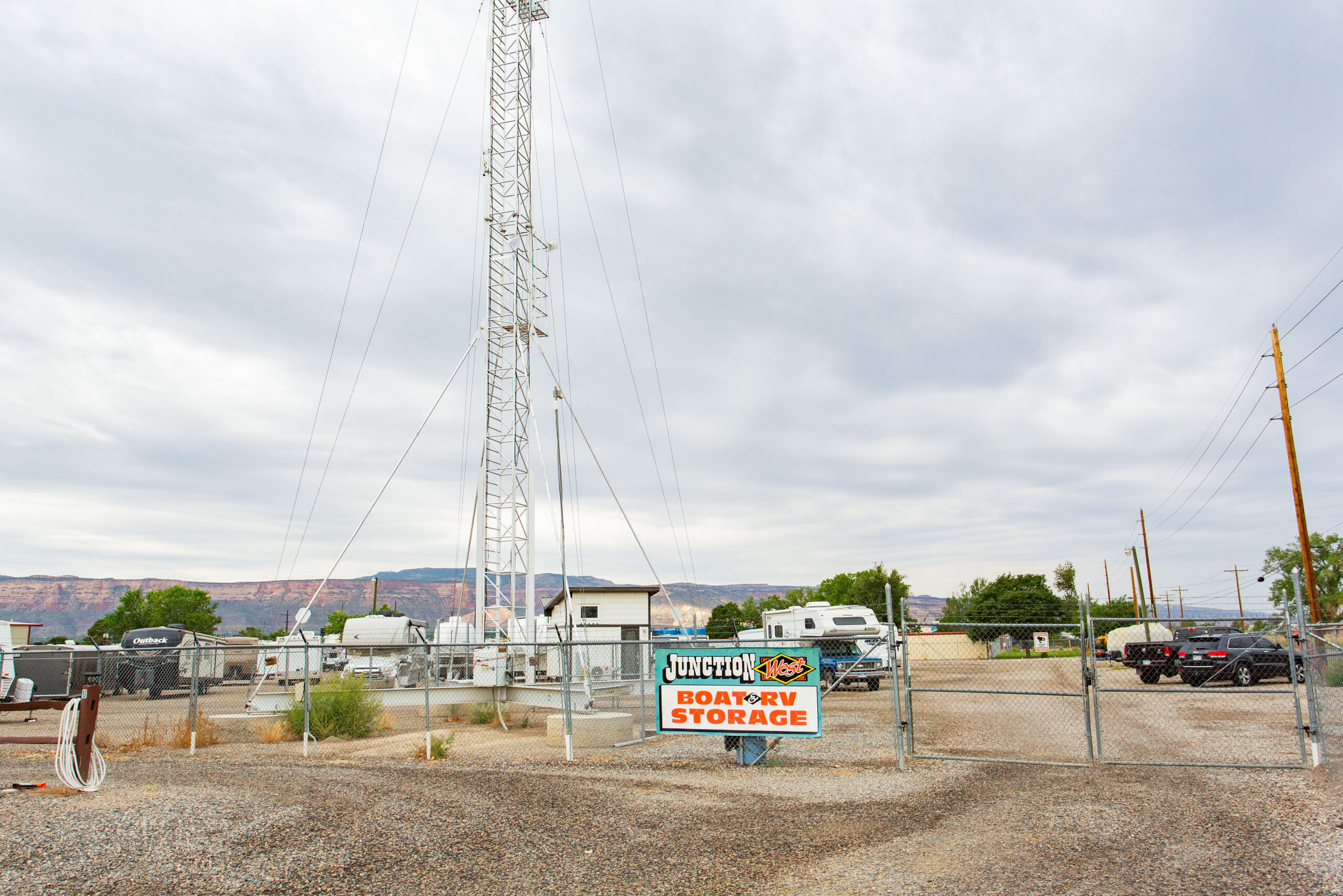 Fenced storage lot with a sign reading “Junction Boat & RV Storage,” several vehicles, RVs, and a tall communication tower in the background under a cloudy sky.