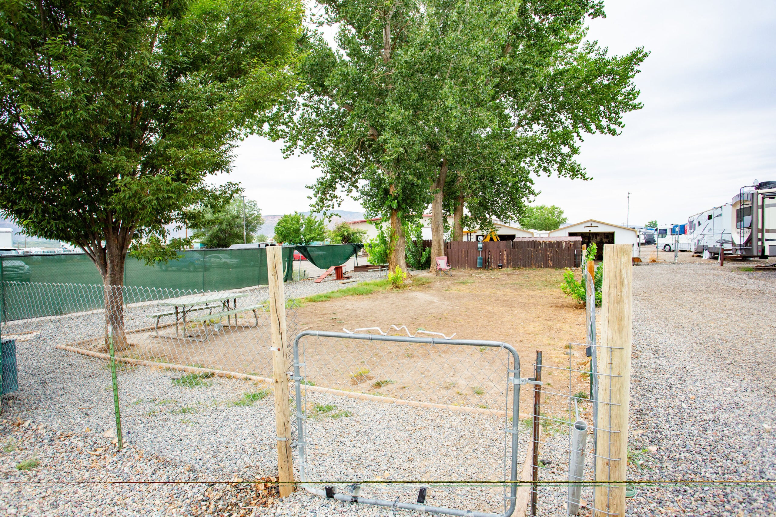 A fenced gravel lot with sparse grass, a few trees, picnic tables, and RVs parked on the right side under an overcast sky.
