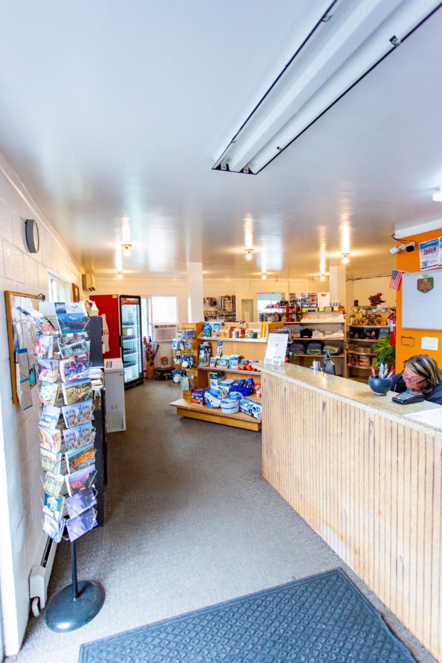 Bright shop interior with a wooden counter, a display rack of postcards, shelves of products, and a person seated behind the counter.