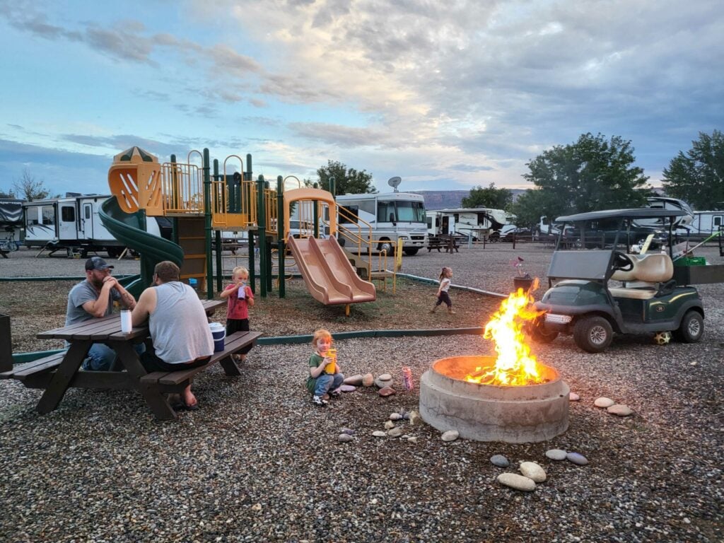 Several people sit at a picnic table near a playground and a campfire, with a golf cart and RVs in the background under a cloudy sky.
