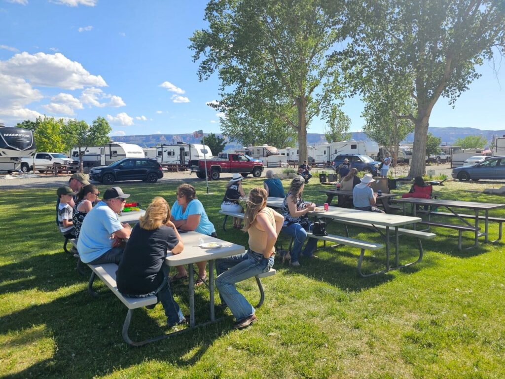 People sit at picnic tables on a grassy area at an RV park, eating and talking under sunny skies with trees and trailers in the background.