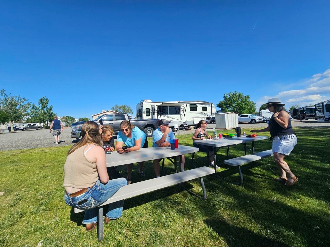 Six people sit and stand around picnic tables in a grassy area at an RV park on a sunny day, with motorhomes and trees visible in the background.