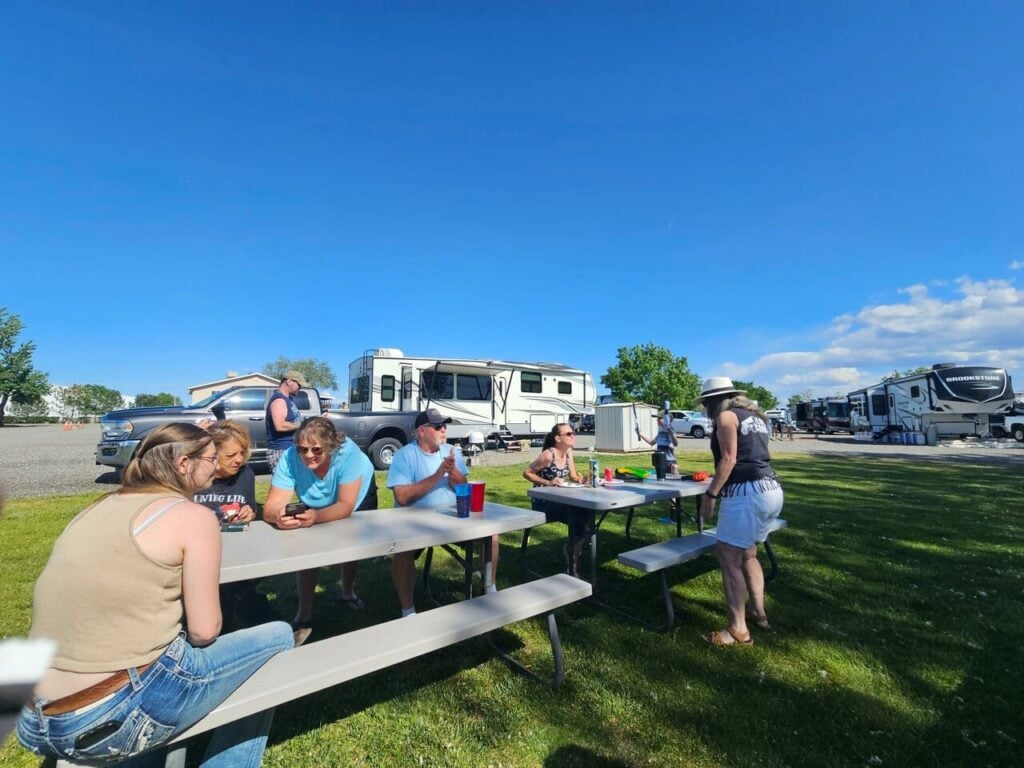 A group of people sit and stand around picnic tables on grass near RVs under a clear blue sky at a campground.