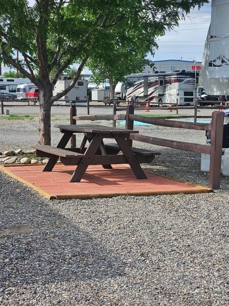 Wooden picnic table and bench set on a red mat under a tree, surrounded by gravel, with RVs and a fence in the background.