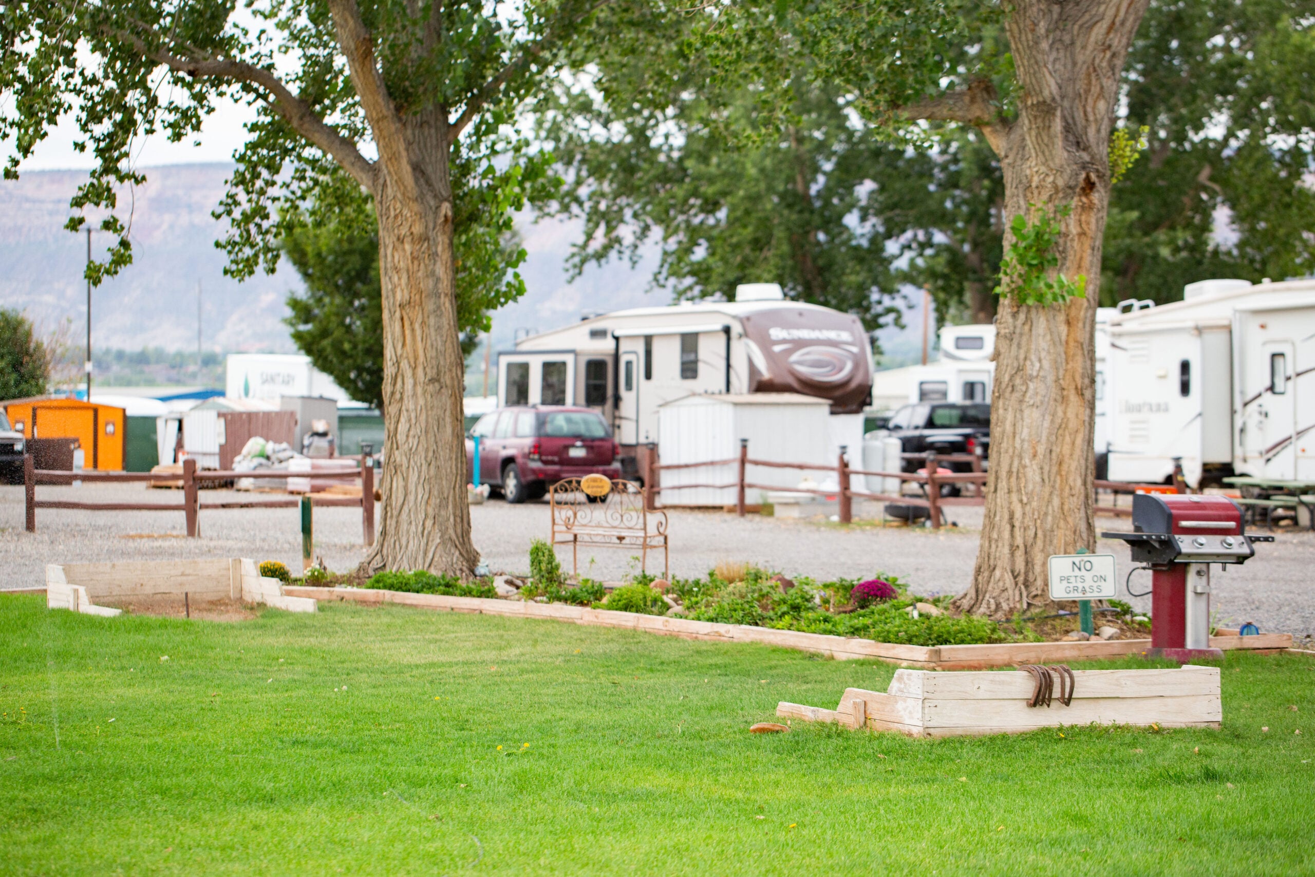 A grassy area with a horseshoe pit, picnic table, sign, grill, and several parked RVs in the background at a campground.