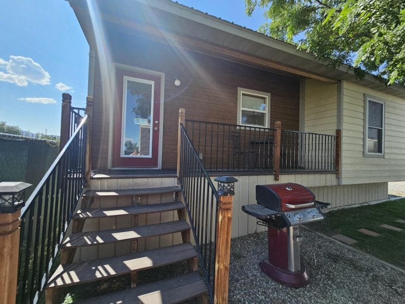 Small house with a wood porch, metal railings, steps, and a red barbecue grill on a gravel area, under a sunny sky with some trees and clouds.