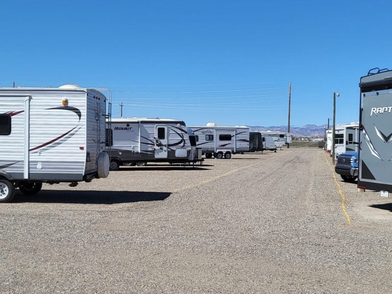 Several RVs are parked in two rows on a gravel lot under a clear blue sky, with mountains visible in the distant background.