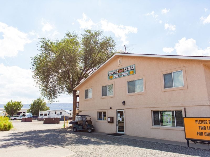 Tan two-story building with a sign reading “Office,” a golf cart parked outside, and an orange sign directing check-in lines; RVs and trees in the background under a bright sky.