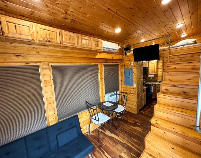 Interior of a small wooden cabin with a black futon, a table with two chairs, a mounted TV, kitchen area, and stairs leading to a loft.
