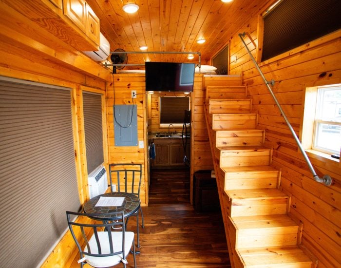 Interior of a tiny house with wooden walls, a small dining table and chairs, stairs leading to a loft, and a view into a compact kitchen.