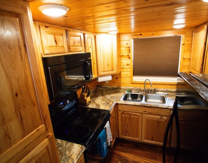 A small kitchen with wood-paneled cabinets, a black stove and microwave, a double sink, granite countertops, and a stainless steel refrigerator.