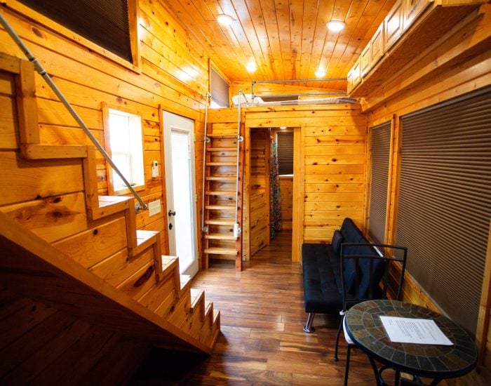 Interior of a small wooden cabin with stairs leading to a loft, a black futon, a round table with papers, and wood-paneled walls and ceiling.