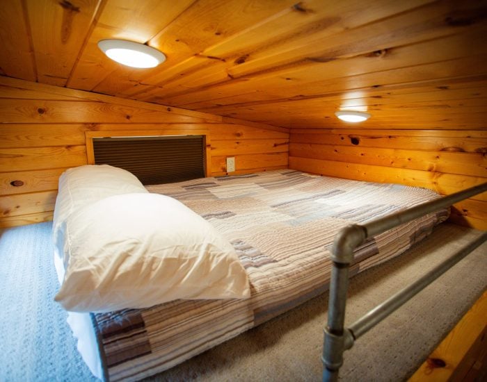A small loft bedroom with a low wooden ceiling, a double bed with striped bedding and two pillows, carpeted floor, and two round ceiling lights.