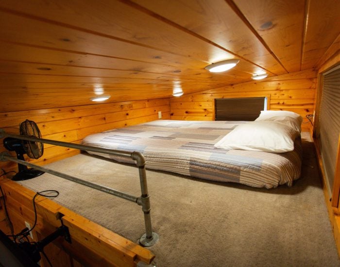 A small loft bedroom with a low wooden ceiling, a bed with striped bedding, two pillows, built-in lights, a fan, and carpeted floor.