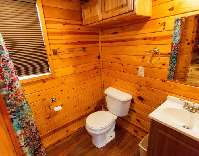 Small bathroom with wood-paneled walls, a white toilet, a sink with a mirror, a window with a blind, and a colorful shower curtain.