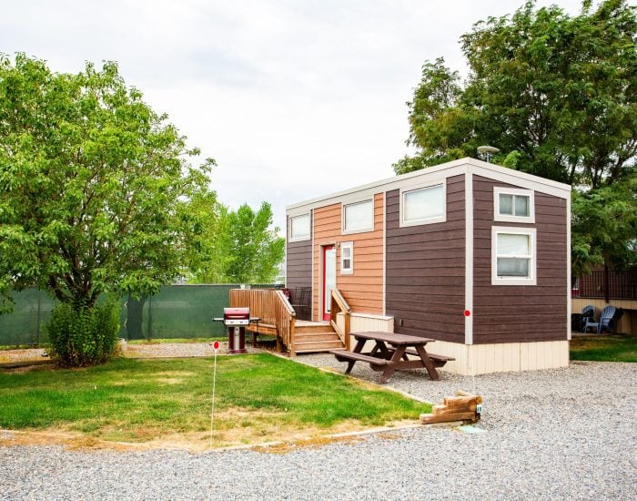 A small, modern tiny house with brown and tan siding sits on a gravel lot, featuring a wooden porch, picnic table, and surrounding trees.