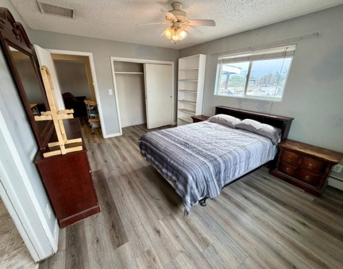 A bedroom with a queen-sized bed, a dark wood dresser with a mirror, a built-in closet, and a white bookshelf on a wood-look floor.