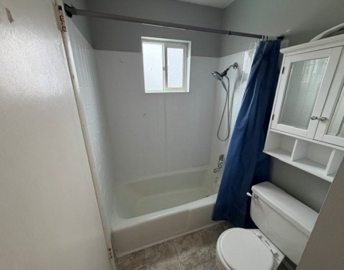 A bathroom with a bathtub, shower head, blue curtain, toilet, and white medicine cabinet.