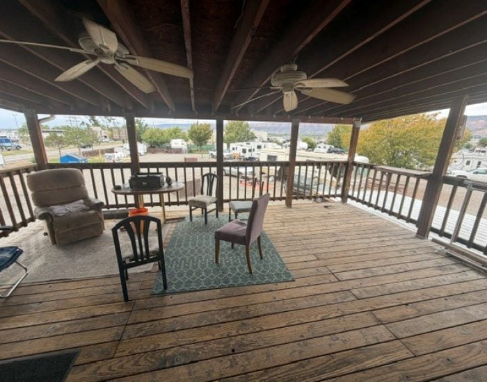 A wooden covered deck furnished with an armchair, four dining chairs, a small table, and a rug, overlooking a parking lot with RVs.