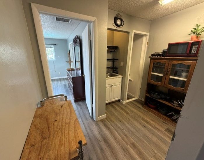 A small kitchen area with a wooden storage hutch, a microwave, and a separate nook containing a sink and cabinets, with a view into a bedroom beyond.
