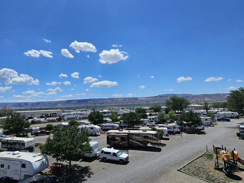 A large RV park with parked trailers and motorhomes on gravel lots under a bright blue sky with scattered clouds.